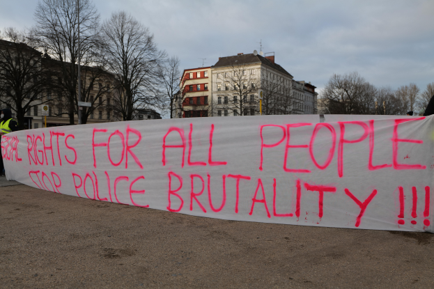Eine Gruppe von Menschen, die auf dem Boden steht und ein Banner hält, auf dem 'Rechte für alle Menschen Stoppt Polizeigewalt' steht, mit einem Straßenpfahl, einem Schild, Bäumen, Gebäuden mit Fenstern und einem bewölkten Himmel im Hintergrund.