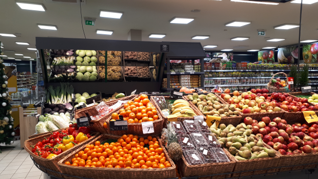 Ein Ladeneinrichtungsbild mit Körbüssern, Obst und Gemüe in Körbchen, Preisschildern, einem dekorierten Weihnachtsbaum links und Regalen mit Artikeln im Hintergrund.