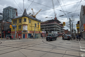 Eine Straßenszene in der Stadt mit einem gelben Gebäude im Bau, Autos, Fußgängern, Strommasten, Ampeln, Gebäuden, Schildern, Bäumen, Kränen und einem bewölkten Himmel.