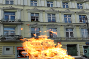 Ein großes Feuer brennt vor einem Gebäude mit Fenstern, Türen, einem Laternenmast und einem Schild mit Text.