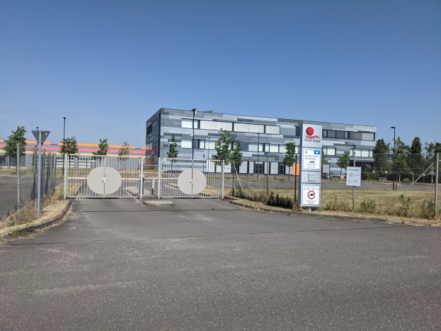 Großes industrielles Gebäude mit der Aufschrift "First Solar" und vielen Fenstern, umgeben von einem Metallzaun, Bäumen, Pflanzen, Gras, Informationsschildern, Straßenmasten und einem Weg unter einem bewölkten Himmel.