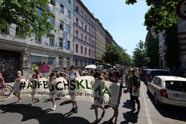 Eine Gruppe von Menschen marschiert mit einem Transparent die Straße entlang, Autos parken rechts und ein Radfahrer links, vor Gebäuden und Bäumen während einer Demonstration in Berlin.