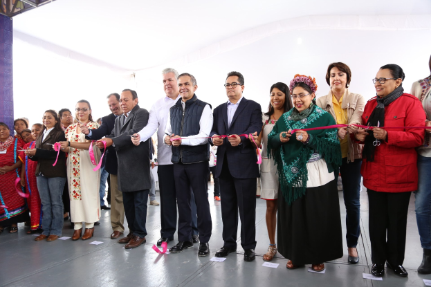 Eine Gruppe von Menschen schneidet einen roten Faden bei einer Zeremonie, lächelt und hält eine Schere in der Hand, mit Papieren auf dem Boden und einem weißen Tuch im Hintergrund.