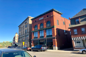 Eine Stadtstraße mit parkenden Autos, historische Gebäude mit Fenstern, Laternen, Schilder, Verkehrszeichen, Bäume und ein Himmel im Hintergrund.