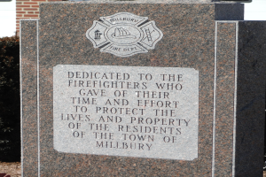 Gedenkstein mit einer Plakette, die den Feuerwehrleuten gewidmet ist, vor einem Backsteingeb├Ąude mit umliegenden Pflanzen und B├Ąumen.