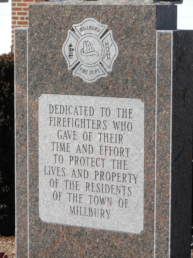 Gedenkstein mit einer Plakette, die den Feuerwehrleuten gewidmet ist, vor einem Backsteingeb├Ąude mit umliegenden Pflanzen und B├Ąumen.