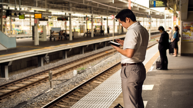 Ein Mann steht auf einem Bahnsteig und schaut auf sein Handy, umgeben von anderen Menschen mit Bahnschienen im Hintergrund; ein Schild mit Text ist auf der rechten Seite sichtbar.