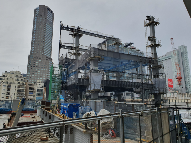 Große Baustelle eines neuen Kraftwerks in einer Stadtumgebung, umgeben von einem Metallzaun, mit Gebäuden, Masten, einem Banner, Fahrzeugen, Drähten und einem bewölktem Himmel.