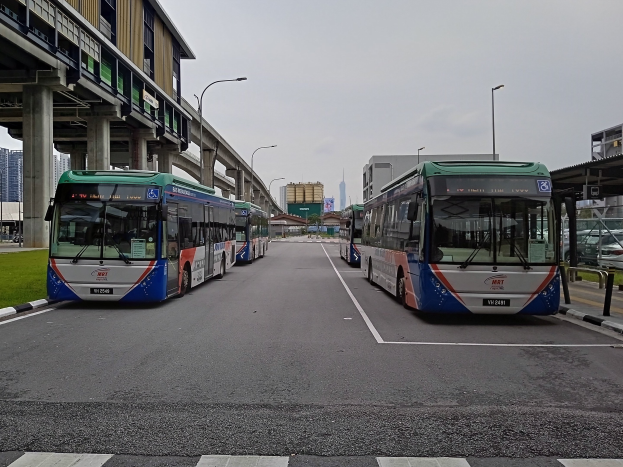 Gruppe von Bussen, die auf der Seite einer Straße geparkt sind, umgeben von Gras, Laternen, einer Brücke mit Säulen, Gebäuden und einem Himmel im Hintergrund.