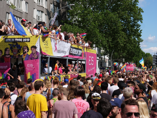 Eine große Menge marschiert auf einer Straße während des Christopher Street Day 2019 in Berlin, viele tragen Mützen und Schutzbrillen, einige halten Fahnen, mit Bannern, Laternenmasten, Bäumen und Gebäuden im Hintergrund unter einem bewölkten Himmel.