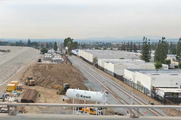 Baustelle mit einer Bahnschiene, Gebäuden, Bäumen, Fahrzeugen, Menschen und einem Geländer, vor Bergen und einem klaren blauen Himmel.