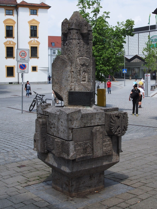 Steindenkmal auf einem Stadtplatz umgeben von Menschen in bunten Kleidern, mit einem Fahrrad daneben und verschiedenen städtischen Elementen im Hintergrund.