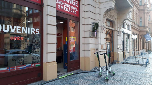Eine Straße in einer Stadt mit parkenden Rollern, Gebäuden, Texttafeln, einem Geländer, Bäumen und einem klaren blauen Himmel.