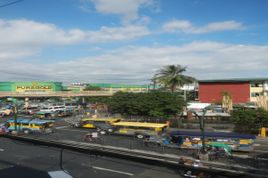 Eine belebte Stadtstraße mit verschiedenen Fahrzeugen, Fußgängern auf Gehwegen, Gebäuden, Bäumen, einem Zaun, Strommasten mit Drähten und einem bewölkten Himmel.