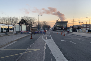 Strassenszene mit Verkehrskegeln, einem Bus, Laternenmästen, Bäumen, Gebäuden, Kränen und Rauch, der aus einem entfernten Gebäude aufsteigt, vor einer Stadtkulisse.