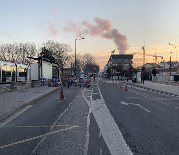 Strassenszene mit Verkehrskegeln, einem Bus, Laternenmästen, Bäumen, Gebäuden, Kränen und Rauch, der aus einem entfernten Gebäude aufsteigt, vor einer Stadtkulisse.