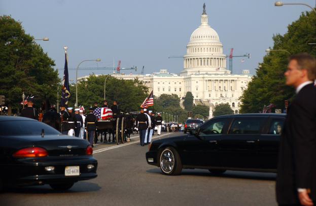 Trauerzug mit Menschen in Mützen und Fahnen vor dem Kapitolsgebäude, Fahrzeuge, Laternenpfähle, Bäume und Gebäude unter einem klaren Himmel.