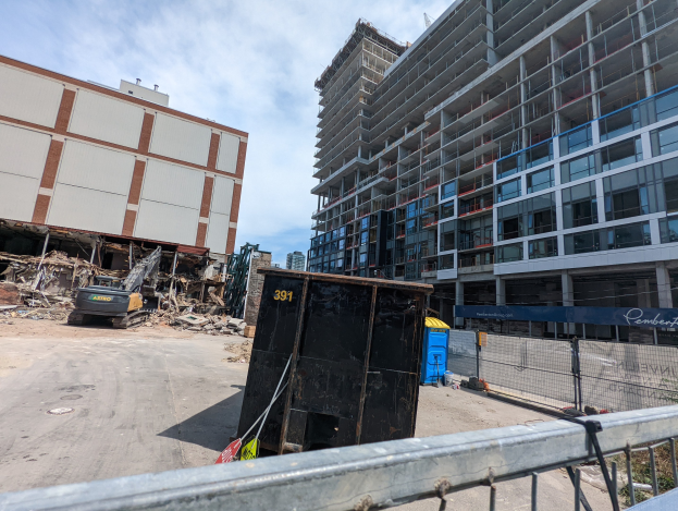 Großes Gebäude mit Schutt davor, umgeben von einem Metallzaun, einem Container, Pflanzen, Bäumen, Pfählen, einem Schild, Fahrzeugen, Steinen und einem bewölkten Himmel an einem Bauplatz für ein neues Wohngebäude.