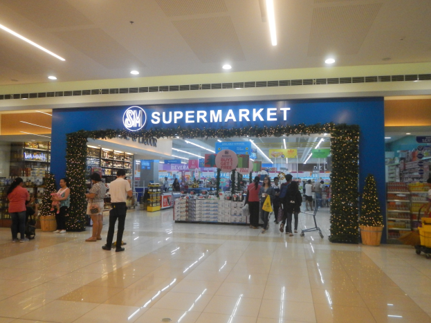 Menschen vor einem Supermarkt mit Taschen, Einkaufswagen, Topfpflanze, Regalen, Schildern und Deckenbeleuchtung.