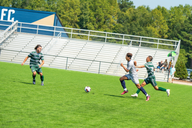 Junge Männer spielen Fußball auf einem Rasenfeld mit Bäumen, einem Schuppen und Zuschauern im Hintergrund unter einem Schirm.