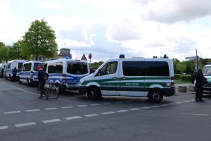 Eine Gruppe von Polizeifahrzeugen, die entlang einer Straße geparkt sind, mit einem Radfahrer im Vordergrund, Bäumen, Polen, Schildern, Gebäuden und einem bewölkten Himmel im Hintergrund.