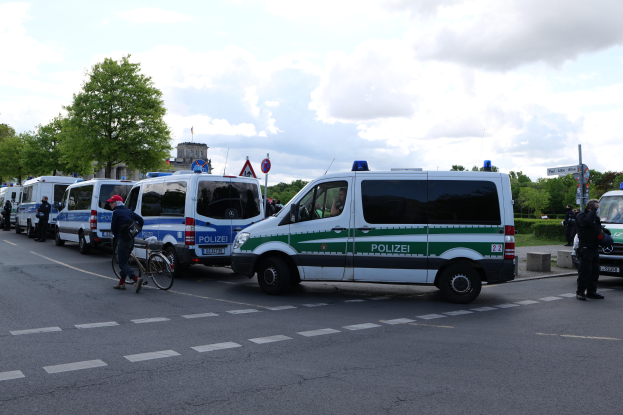 Eine Gruppe von Polizeifahrzeugen, die entlang einer Straße geparkt sind, mit einem Radfahrer im Vordergrund, Bäumen, Polen, Schildern, Gebäuden und einem bewölkten Himmel im Hintergrund.