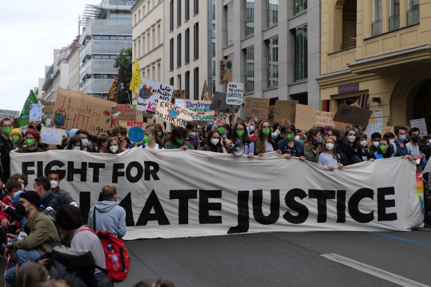 Eine große Gruppe von Menschen marschiert mit einem Banner, auf dem "Kampf für Hassjustiz" steht, durch eine Straße, einige tragen Masken und halten Schilder oder Taschen, mit Gebäuden, Bäumen und einem klaren blauen Himmel im Hintergrund.