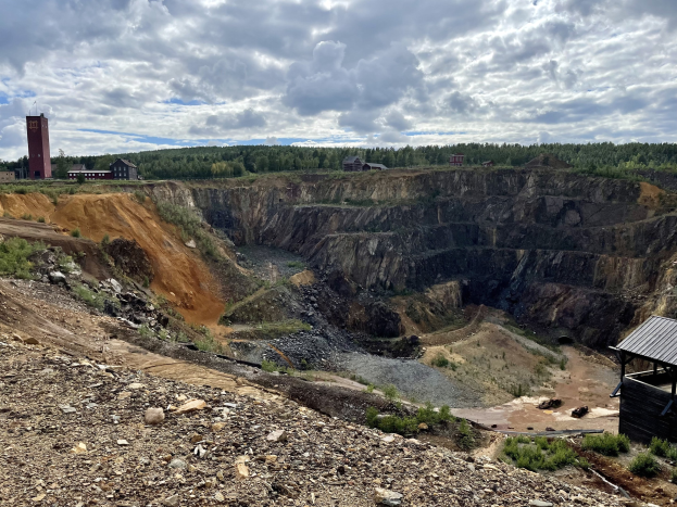Großer Tagebau umgeben von Bäumen, Gebäuden und einem Schuppen, mit bewölktem Himmel und steinigem, bepflanztem Boden.