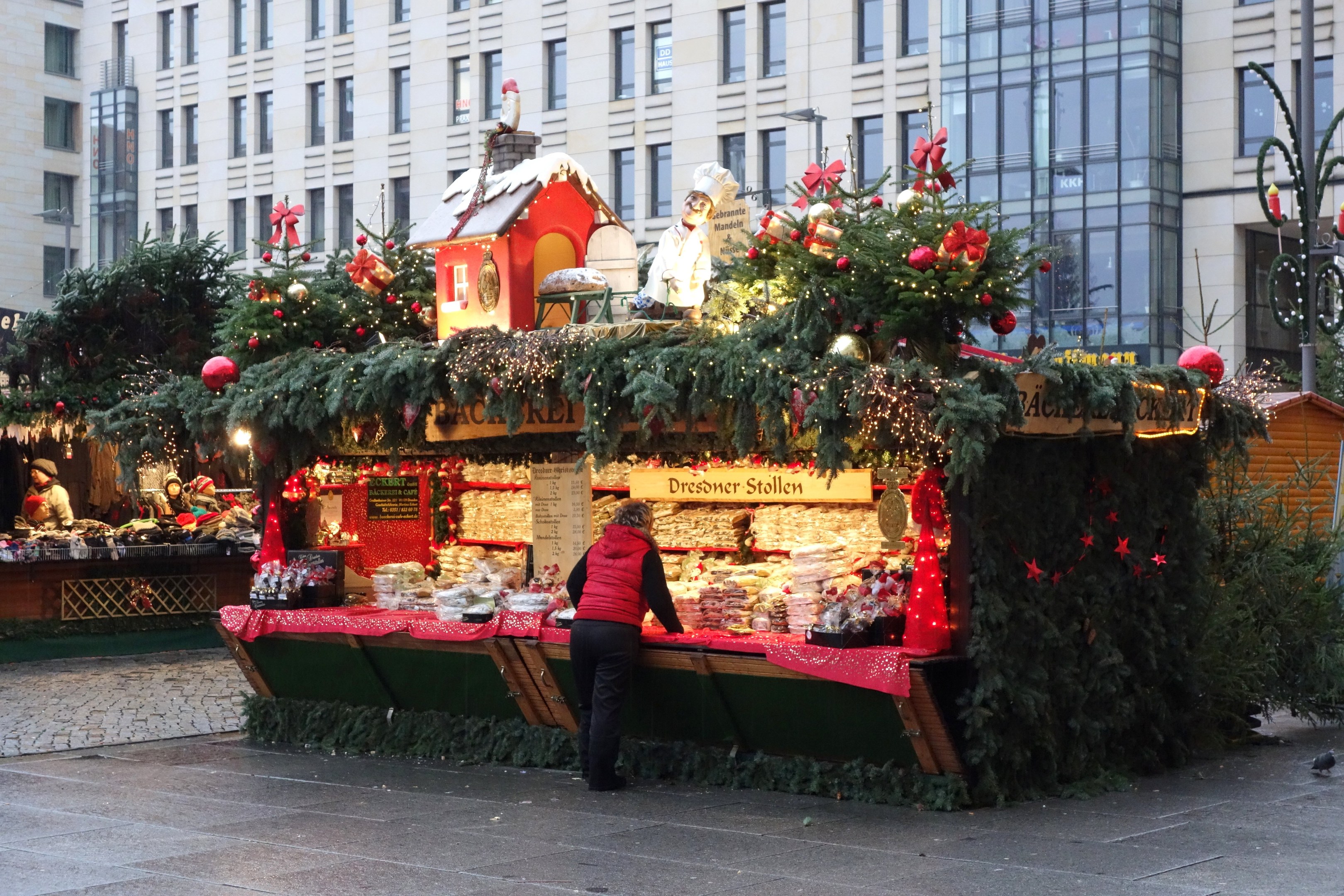 Eine Person steht vor einem Weihnachtsmarktstand auf einem Stadtplatz, umgeben von festlichen Dekorationen und anderen Menschen, mit Gebäuden und einem Laternenmast im Hintergrund.