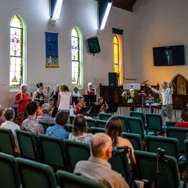 Eine Gruppe von Menschen sitzt auf Stühlen in einer Kirche, ein Mann steht vorn mit einem Mikrofon, umgeben von Musikinstrumenten, einem Tisch mit einer Blumenvase, einem Lautsprecher, einer Fahne mit Text, Fenstern, einem Fernseher und Deckenleuchten.