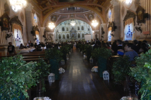 Eine Hochzeitszeremonie in einer Kirche mit Gästen auf Bänken sitzend und im Gang stehend, dekoriert mit Zimmerpflanzen, Skulpturen, Vorhängen, Kronleuchtern und einem Bildschirm im Hintergrund.