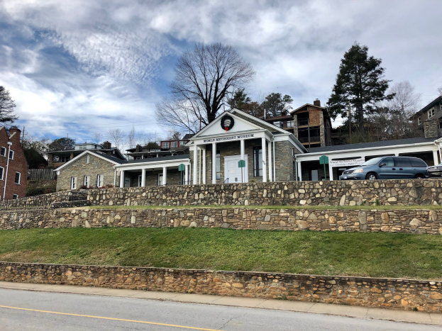 Steinwand mit parkenden Autos, benachbarten Gebäuden mit Fenstern und Türen, Bäumen und einem bewölkten Himmel im Hintergrund, als Weltkulturerbe-Museum etikettiert.
