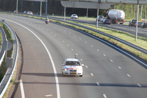 Ein Auto fährt auf einer Autobahn mit einem Polizeiwagen am Straßenrand, anderen Fahrzeugen, Leitplanken, Pfosten, Planken und Bäumen im Hintergrund.