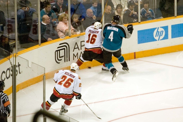 Ein Hockey-Spieler in einem weißen und schwarzen Trikot mit der Nummer 16, Helm, Shorts und Schuhen steht neben einem Mann in einer blauen Jacke, Brille, Helm und Hose neben einem Fenster. Zuschauer sitzen auf Stühlen oben, während ein Spieler im Trikot mit der Nummer 26 einen Stock hält. Ein Schiedsrichter steht hinter Glas links unten und ein Werbeplakat einer Firma ist rechts zu sehen.