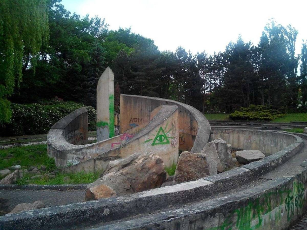 Abandonierter Skatepark mit Graffiti, umgeben von Felsen, Gras, Pflanzen und Bäumen unter einem klaren blauen Himmel.