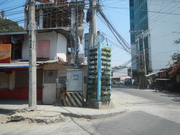 Stadtstraße mit Gebäuden, Strommasten, Kabeln, Schildern, Fahrzeugen, Bäumen und einem Gebäude mit Drähten im Vordergrund.