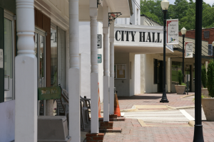 Ein Rathaus umgeben von Gebäuden, Straßenlaternen, Straßenschildern, Autos, Verkehrskegeln, Stühlen, Topfpflanzen, Bäumen und einem bewölkten Himmel.