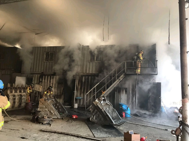 Feuerwehrleute mit Helmen arbeiten daran, ein Lagerfeuer zu löschen, mit Rauch, der aus dem Gebäude aufsteigt, Treppen mit Geländern, die zum Eingang führen, eine Kette an einem Pfahl rechts, eine verglaste Wand im Hintergrund und verstreute Trümmer, einschließlich Kartons und Rohre auf dem Boden.