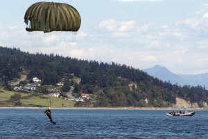 Eine Person segelt über einen See mit einem Boot auf der rechten Seite, Bäume, Gebäude, Hügel und einen klaren blauen Himmel im Hintergrund.