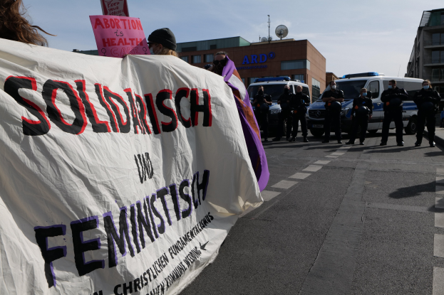 Eine Gruppe von Menschen marschiert mit einem Transparent, auf dem 'Solidarität und Feminismus' steht, vorbei an parkenden Fahrzeugen, Gebäuden, einer Schüsselantenne und einem klaren blauen Himmel.