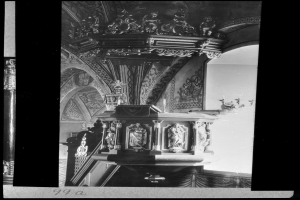 Ein Schwarz-Weiß-Foto eines Kirchenpults mit geschnitzten Säulen und einer strukturierten Wand im Hintergrund, mit Text unten.