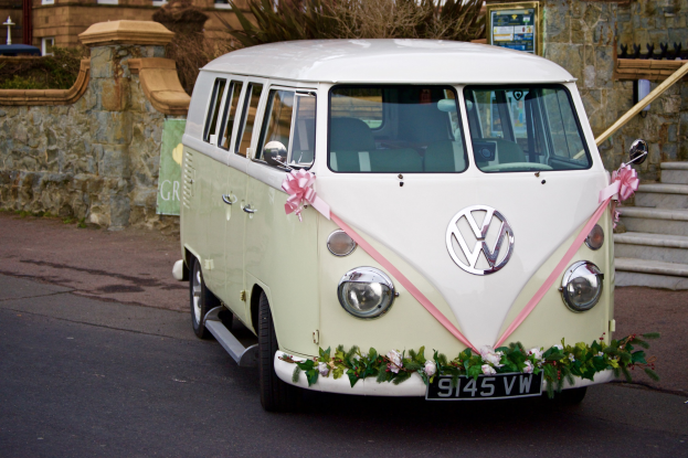 Ein weißer und pinker VW-Campervan, der mit Blumen und Blättern geschmückt ist und vor einer Steinwand mit einer Treppe auf der rechten Seite und einem Gebäude im Hintergrund geparkt ist.