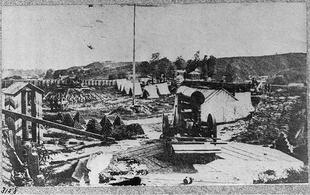 Schwarz-weißes Foto einer Baustelle mit einer Maschine im Vordergrund, umgeben von Hütten, Bäumen, Hügeln und Himmel im Hintergrund, und Text unten, der besagt: "Der Bau einer neuen Brücke in der Stadt San Francisco, Kalifornien."