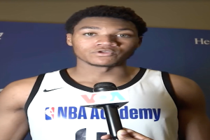 Ein Mann in einem weißen Tanktop hält ein Mikrofon und steht vor einem Banner mit der Aufschrift "NBA All-Star Game 2019" und einer Wand, wahrscheinlich spricht er mit der Medien über das bevorstehende Spiel.