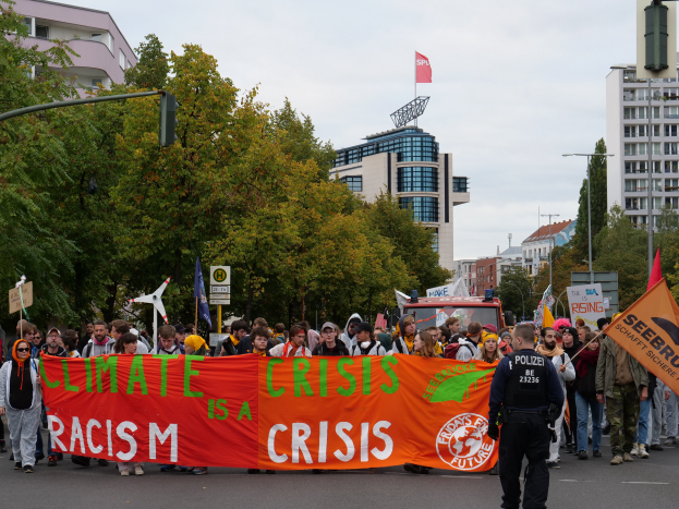 Eine Gruppe von Menschen marschiert eine baumbestandene Straße entlang und hält ein Banner in die Höhe, auf dem 'Klimakrise ist eine Krise' steht, mit Gebäuden und einem klaren blauen Himmel im Hintergrund.