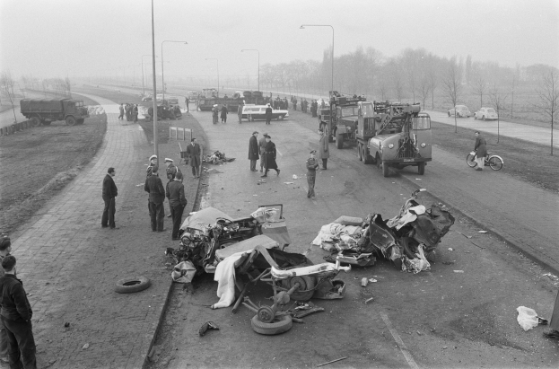 Schwarz-weiß-Szene eines Autounfalls am Straßenrand mit mehreren Fahrzeugen, einer Gruppe von Menschen in der Nähe, Laternenpfählen, Bäumen und dem Himmel im Hintergrund.