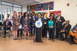 Gruppe von Menschen vor einem Podium mit Mikrofon und einer Tafel mit der Aufschrift "Sancho-Stadtrat tritt dem Stadtrat bei", mit sitzenden und stehenden Teilnehmern, Flaggen, Feuerlöscher und Deckenbeleuchtung im Hintergrund.
