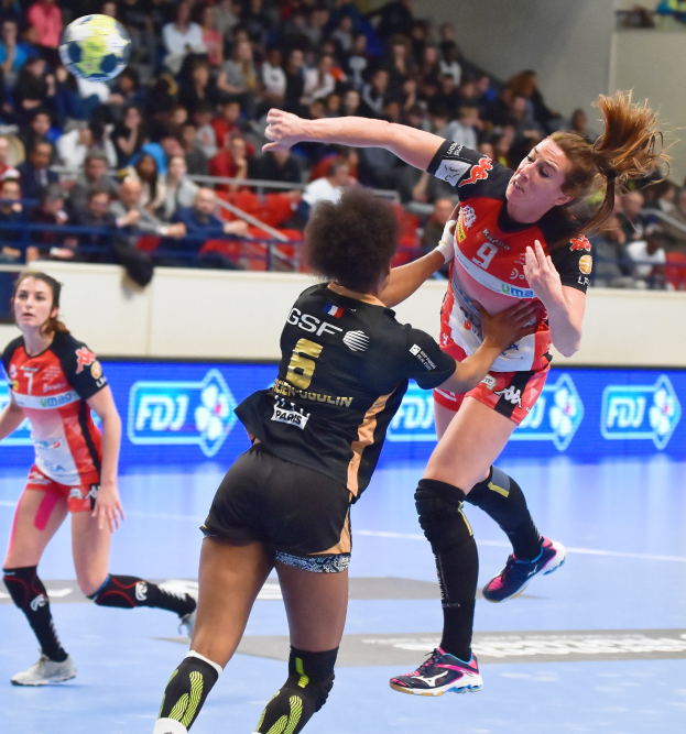Eine Gruppe von Frauen, die auf einem Platz Handball spielen, mit einem Ball in der Luft, beobachtet von Zuschauern auf einer Tribüne.