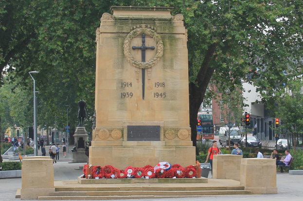 Kriegsdenkmal in einer Stadtstraße umgeben von Kränzen, mit Menschen auf Bänken, Fahrzeugen, Verkehrszeichen, Laternenpfählen, Bäumen, Gebäuden und Himmel im Hintergrund.