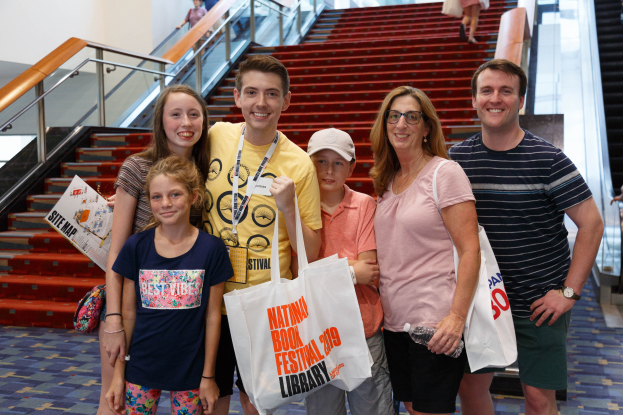 Eine Gruppe von Menschen, die zusammensteht, lächelt und Taschen hält, vor einer Rolltreppe auf der Nationalen Buchmesse 2019, mit Treppen, Geländern und einer Wand im Hintergrund.
